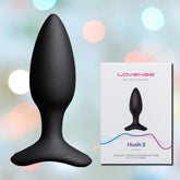 A Lovense Hush 2 Vibrating Butt Plug - Small is displayed next to its packaging. The product and box exhibit a sleek, modern design in black. The packaging highlights Bluetooth remote control, size dimensions, and guarantees intense vibrations along with comfortable wear. The background features a colorful and blurred effect.